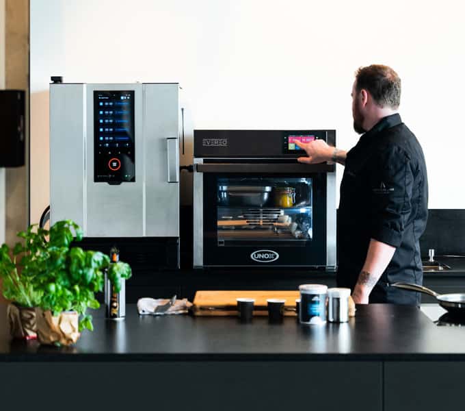 Male chef touches unox evereo control panel, ready meals inside. pan, basil on counter, unox speedx beside evereo.