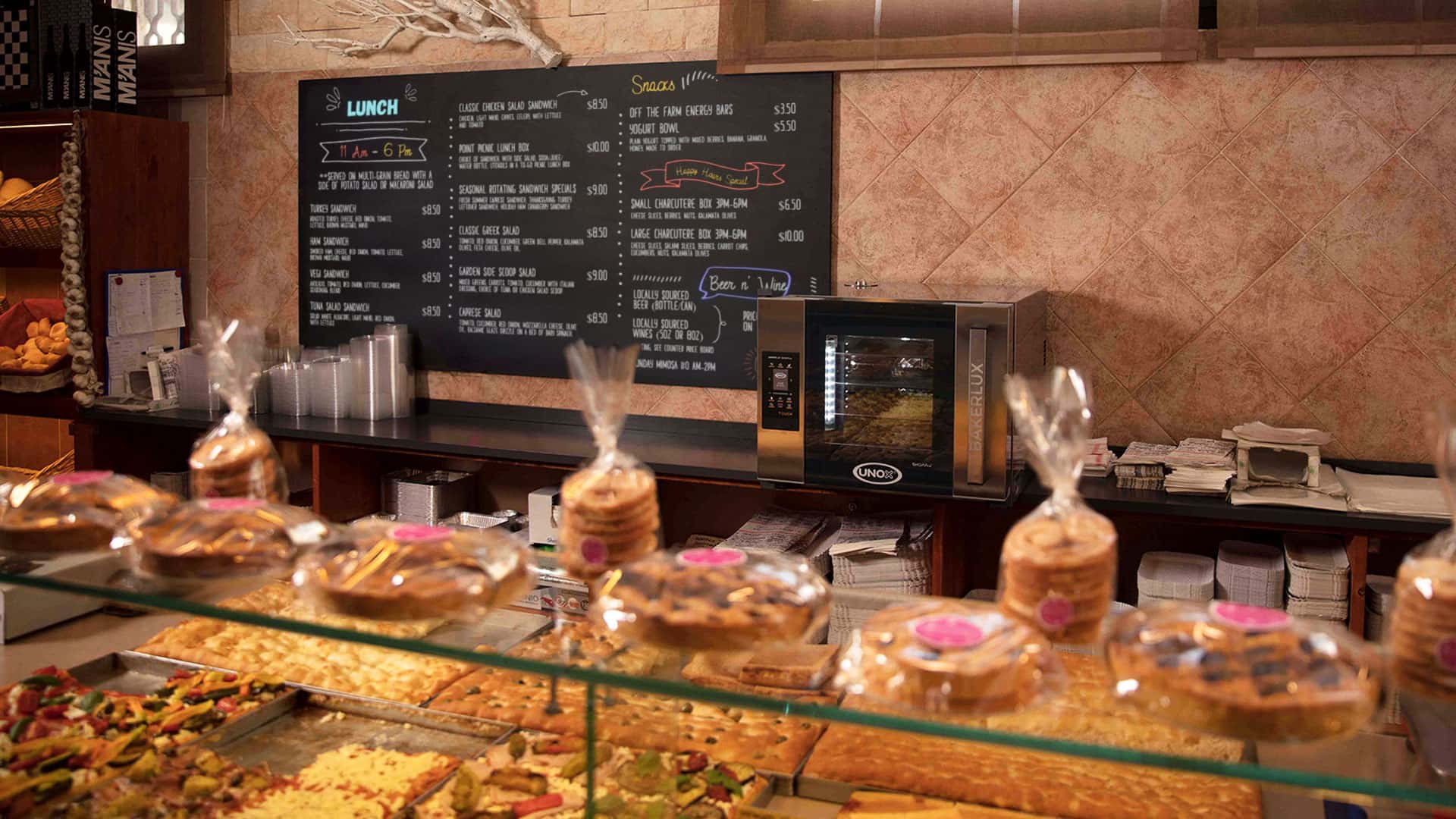 An Unox oven sits on a deli counter. There is a black chalkboard sign on the wall showing the menu. Pastries and foccaccia on the display counter.