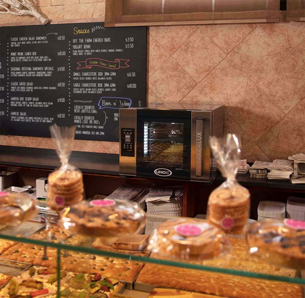 An Unox oven sits on a deli counter. There is a black chalkboard sign on the wall showing the menu. Pastries and foccaccia on the display counter.