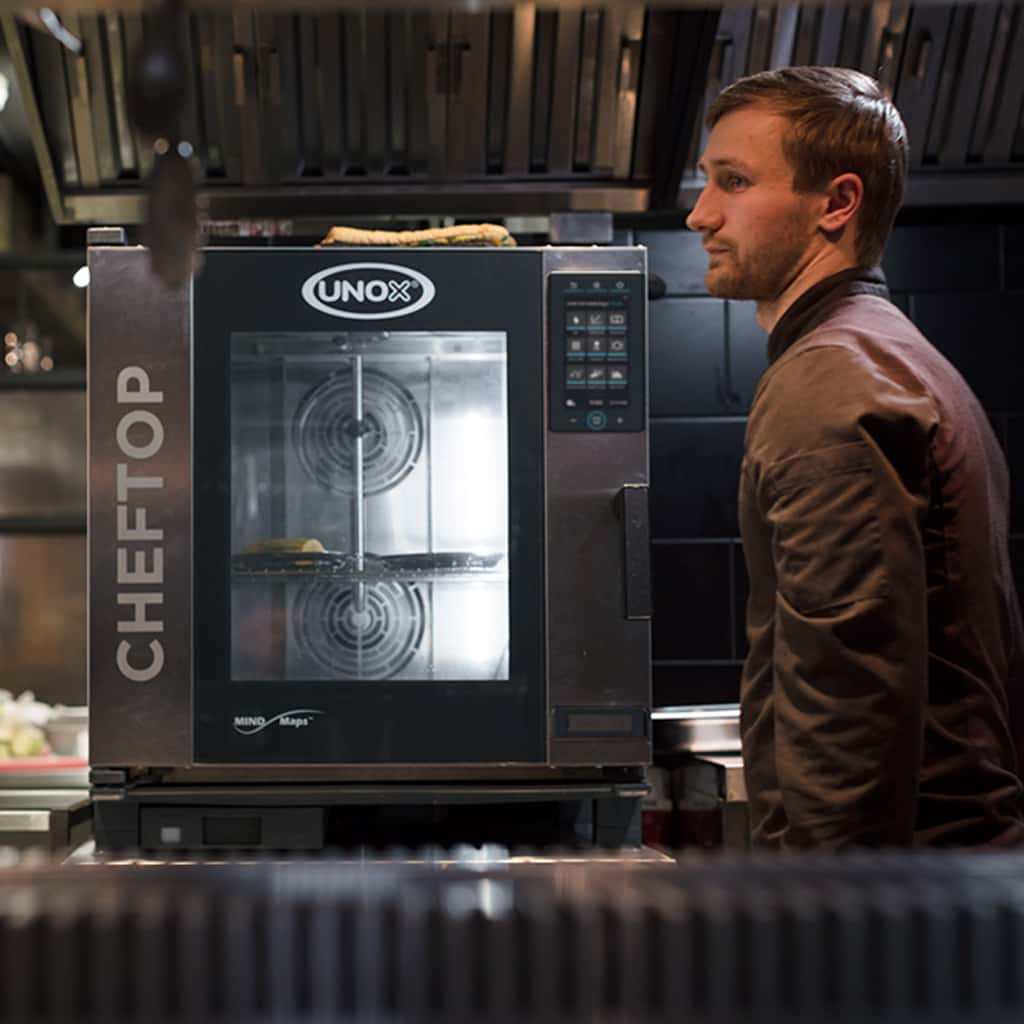 Chef uomo accanto a forno professionale combinato Unox CHEFTOP MINMaps in cucina professionale, con cibo all'interno. 