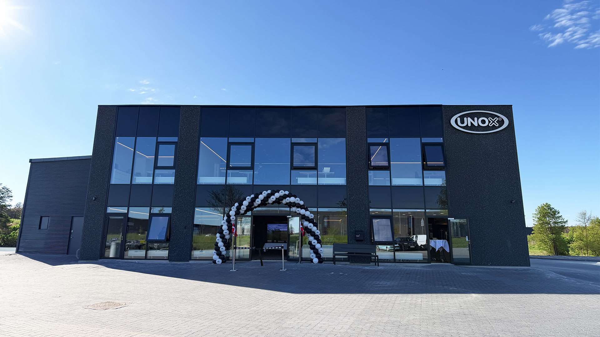 Modern two-story Denmark Experience Center with large windows, a black and white balloon arch at the entrance, and a logo on the facade. Bright, sunny day.