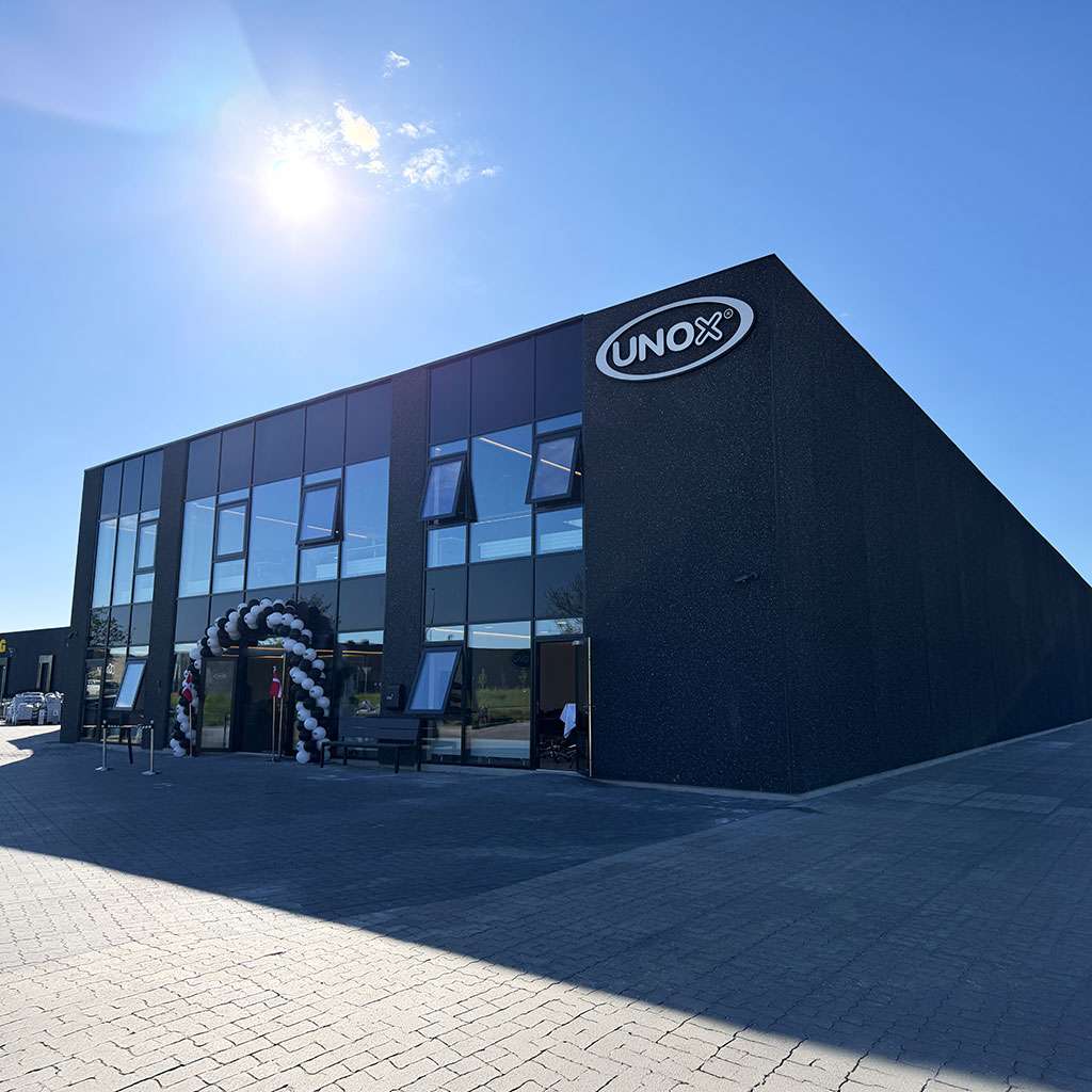 Modern two-story Denmark Experience Center with large windows, a black and white balloon arch at the entrance, and a logo on the facade. Bright, sunny day.