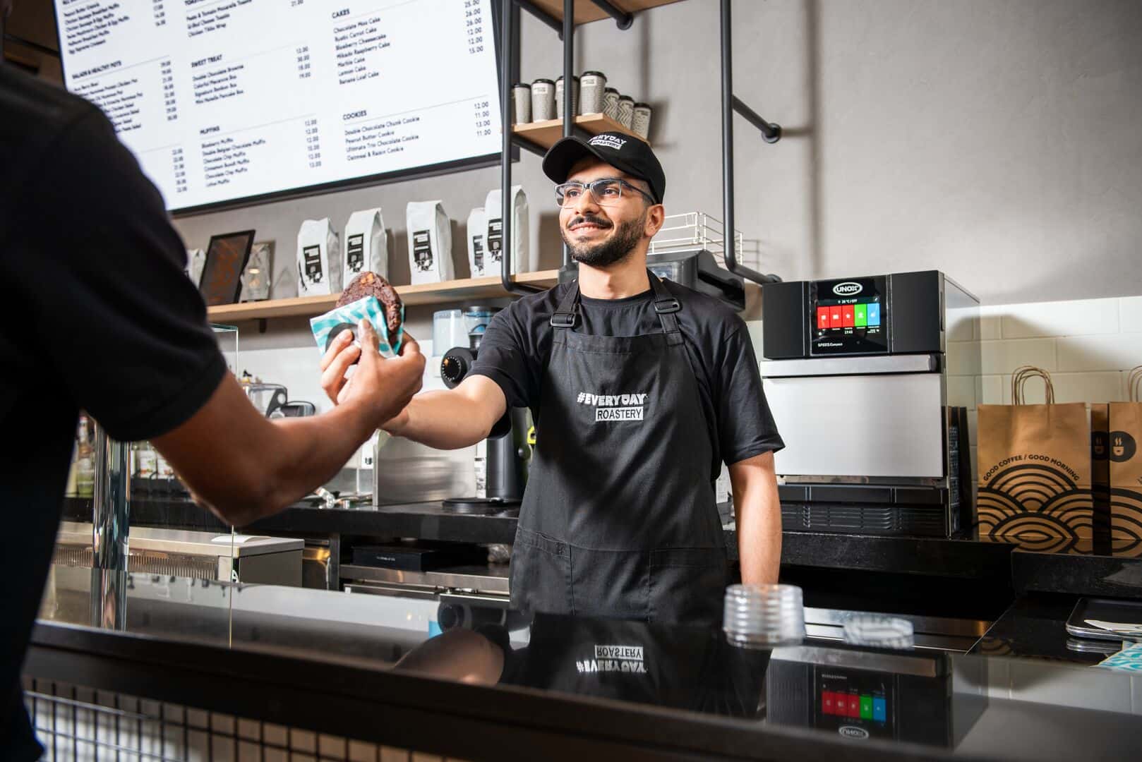 Un barista serve un biscotto caldo a un cliente, con un forno a cottura accelerata compatto Unox visibile sul banco.